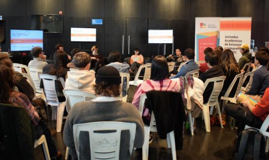 Fotografía del hall de la FIC durante la mesa temática de cierre del evento, encabezada por estudiantes. El espacio está colmado de personas en su mayoría jóvenes sentadas en sillas blancas. El auditorio se ve de espaldas. Los expositores son tres jovenes, dos mujeres y un varón, sentados en sillones negros. Detrás se ven tres pantallas y un roll up que en letras negras sobre fondo blanco con rayas en tonos de color naranja en la parte superior e inferior de la lona dice "IV Jornadas de Extensión".
