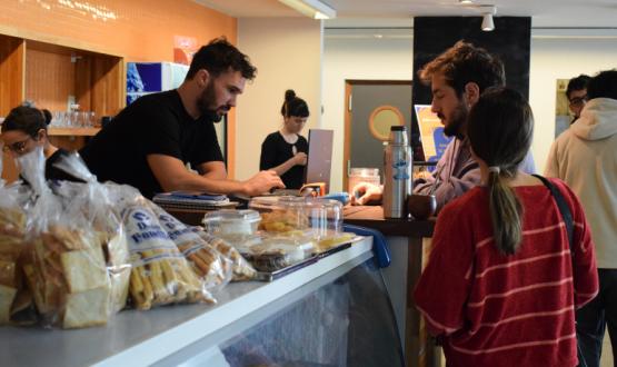 A un lado de un mostrador un hombre sostiene una tarjeta, del otro lado otro hombre digita en un pos. Sobre el mostrador se ve un termo y un mate apoyado y al costado una vitrina con artículos de panadería. 