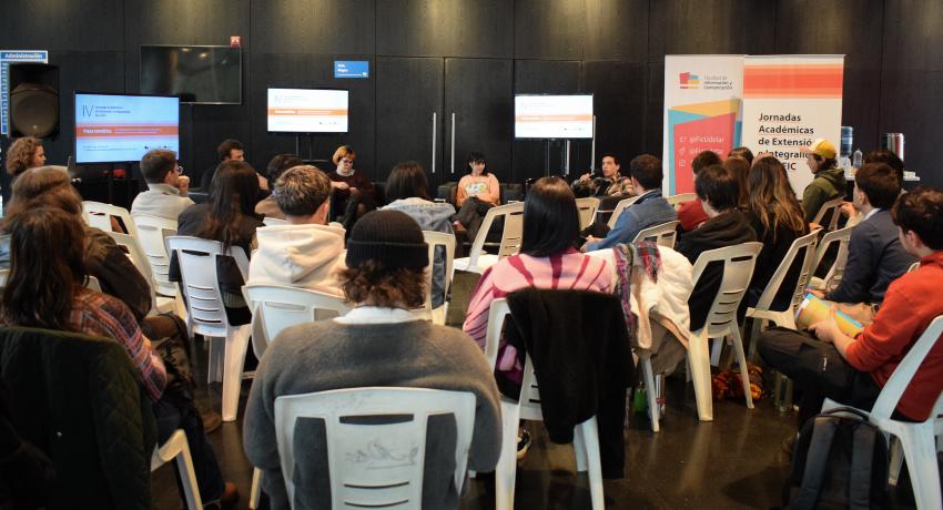 Fotografía del hall de la FIC durante la mesa temática de cierre del evento, encabezada por estudiantes. El espacio está colmado de personas en su mayoría jóvenes sentadas en sillas blancas. El auditorio se ve de espaldas. Los expositores son tres jovenes, dos mujeres y un varón, sentados en sillones negros. Detrás se ven tres pantallas y un roll up que en letras negras sobre fondo blanco con rayas en tonos de color naranja en la parte superior e inferior de la lona dice "IV Jornadas de Extensión".