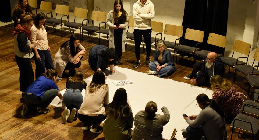 Fotografía. En un salón, un grupo de personas trabajan en torno a un papelógrafo apoyado en el piso. Algunas están paradas, otras sentadas en el suelo y otras de rodillas. Algunas tienen marcadores y escriben en el papel.