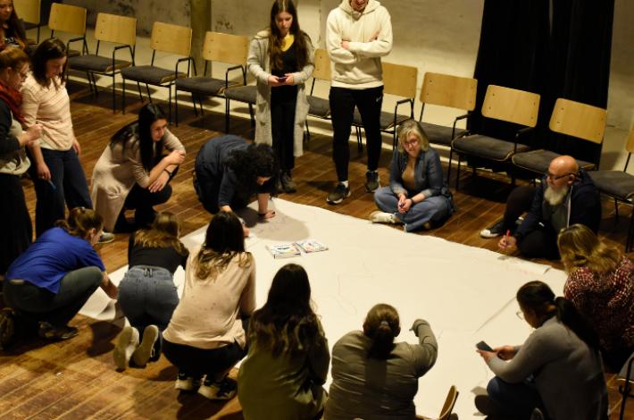 Fotografía. En un salón, un grupo de personas trabajan en torno a un papelógrafo apoyado en el piso. Algunas están paradas, otras sentadas en el suelo y otras de rodillas. Algunas tienen marcadores y escriben en el papel. 