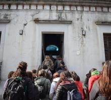 Fotográfía de los estudiantes de espaldas, subiendo la escalinata de la entrada del Museo. A los costados de la puerta, dos ventanas con postigones antiguos marrones cerrados.  