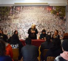 Foto de la sala de conferencias del MUME. La directora del Museo Begoña Ojeda parada detrás de una mesa con mantel rojo se dirige a los visitantes que están sentados en sillas. Detrás de Ojeda, una foto aérea de una de las marchas del 20 de mayo. 