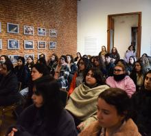 La sala de conferencias está llena de visitantes, qeu están sentados. Fotos de las marchas del silencio entre tres filas cubren una de las paredes del salón con ladrillo a la vista. 