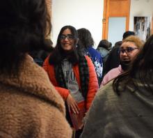 Foto. Sala de exposiciones. Una mujer de pelo largo castaño oscuro y campera roja sonrie. Otra de pelo claro atado y lentes de marco azul mira con gesto de asombro. 