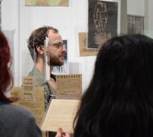 Foto. Sala de exposiciones del museo. De perfil Un estudiante pelirojo, de barba y lentes de marco grueso mira recortes de diario en soportes transparentes que cuelgan del techo por toda la habitación. 