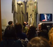 Foto. Sala de exposiciones del Museo. Varios estudiantes observan uniformes carcelarios de color verde que se exponen, colgados del techo de la habitación. 