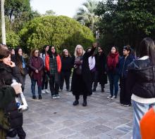 Foto. Afuera del Museo la directora Begoña Ojeda con un grupo de estudiantes que la rodea señala con el brazo estirado hacia lo alto. Detrás hay árboles frondosos. 