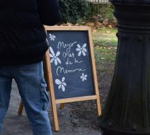 Una pizarra de pie con marco de madrea en el parque exterior del museo. en letras amarillas sobre fondo negro dice "Mayo mes de la memoria" y cuatro margaritas dibujadas con tiza blanca. hay un hombre mirando el cartel, de espaldas, lleva jean y campera negra. 